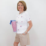 Woman wearing a white polo shirt with logos and beige shorts, holding a colorful bag against a white background.