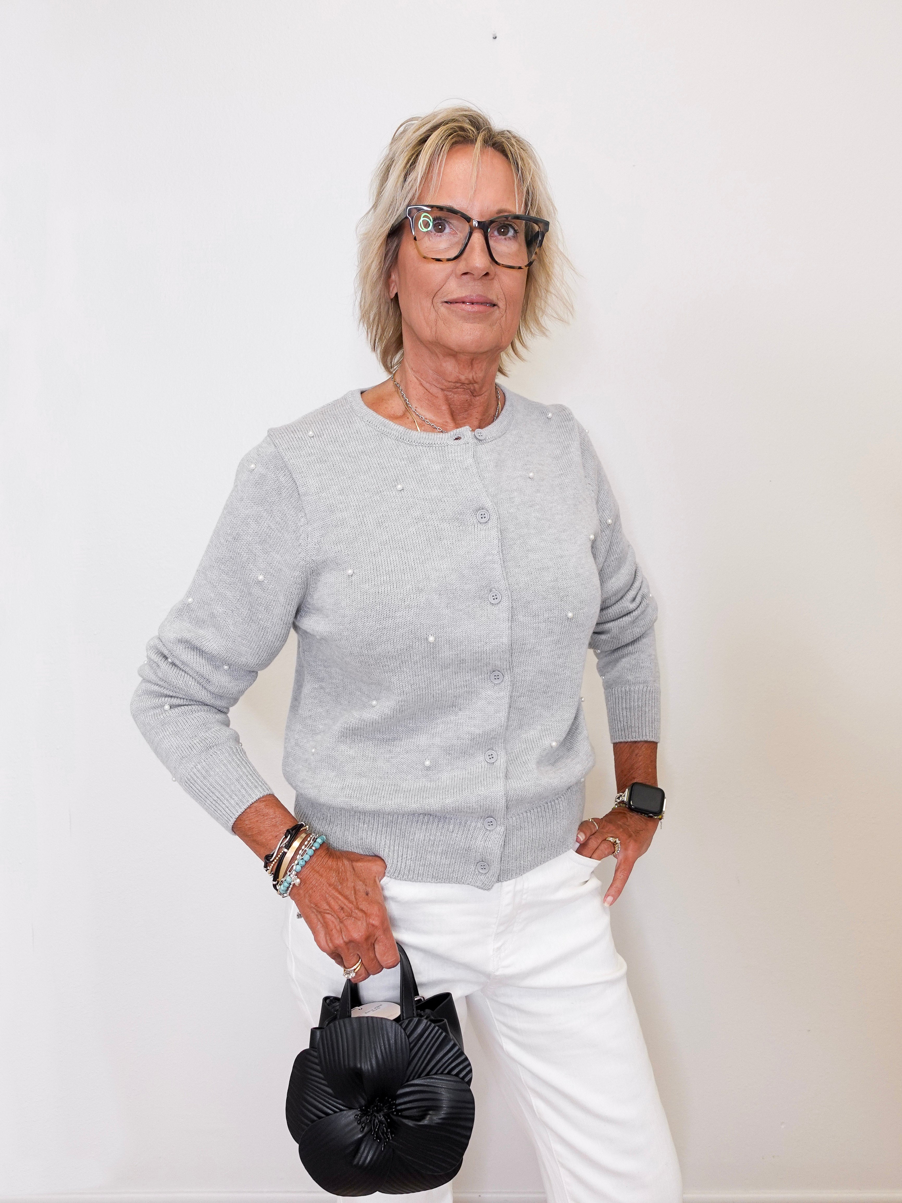 Woman wearing a gray sweater and white pants holding a black handbag against a white background