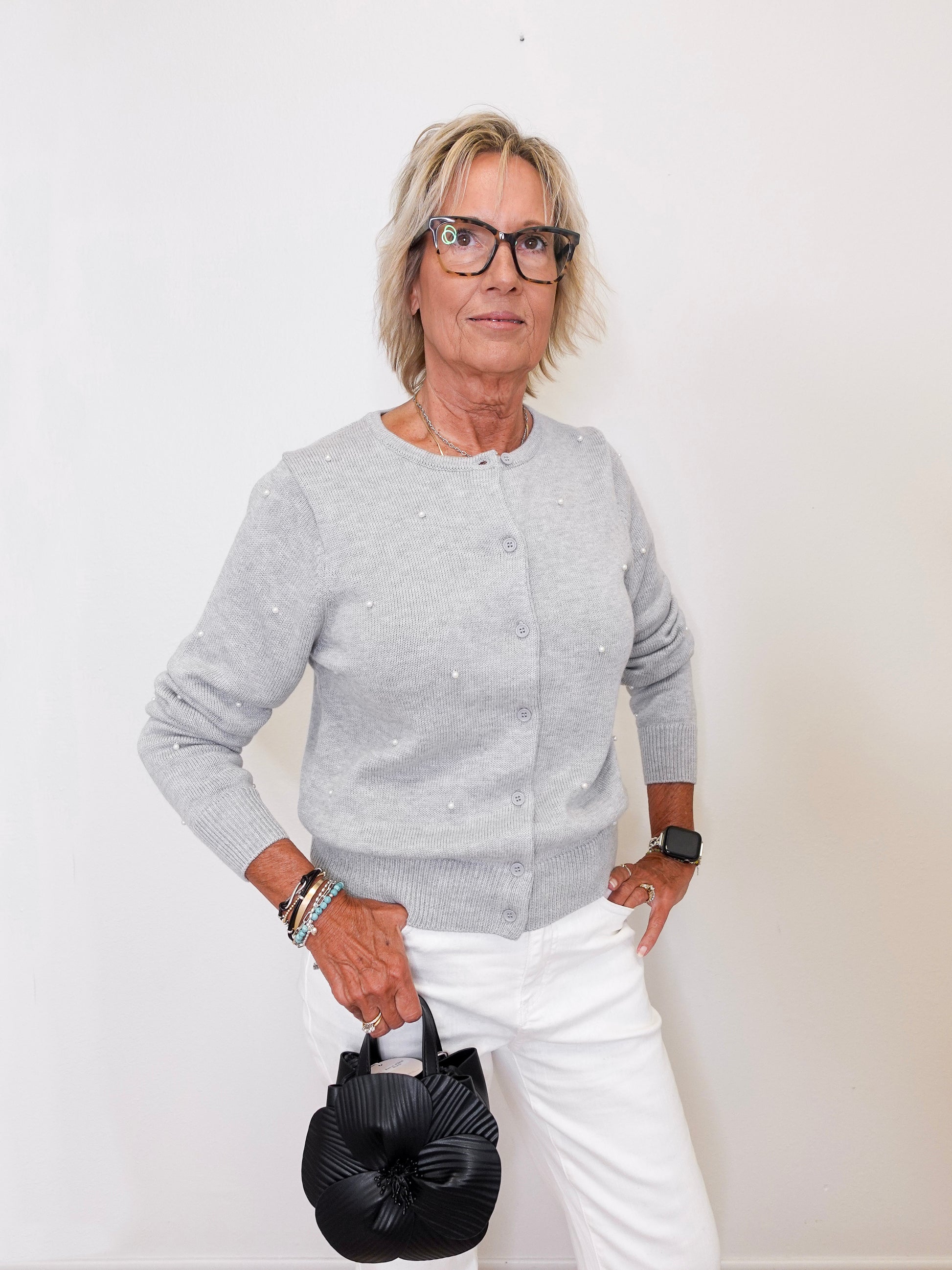Woman wearing a gray sweater and white pants holding a black handbag against a white background