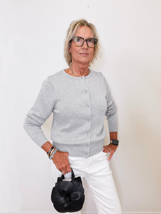 Woman wearing a gray sweater and white pants holding a black handbag against a white background