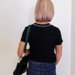 Person wearing a black t-shirt with decorative elements and blue jeans, holding a black bag against a white wall.