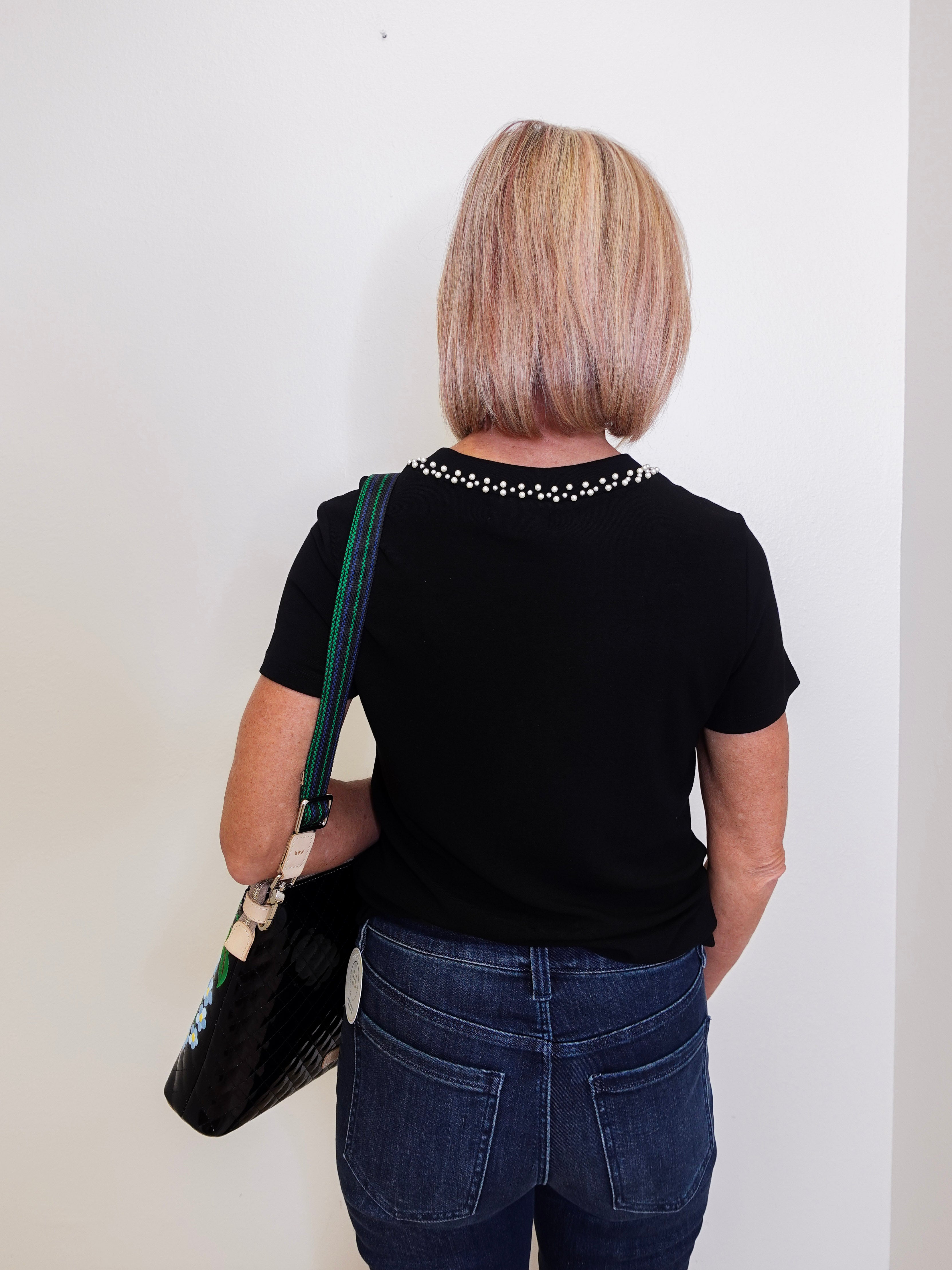 Person wearing a black t-shirt with decorative elements and blue jeans, holding a black bag against a white wall.