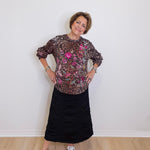 Woman wearing a patterned top and black skirt standing against a white wall.