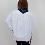 A woman viewed from behind wearing a white long-sleeve button-up shirt with a black neck scarf featuring a blue geometric pattern, paired with black pants and holding a small black handbag.
