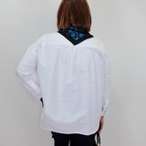 A woman viewed from behind wearing a white long-sleeve button-up shirt with a black neck scarf featuring a blue geometric pattern, paired with black pants and holding a small black handbag.