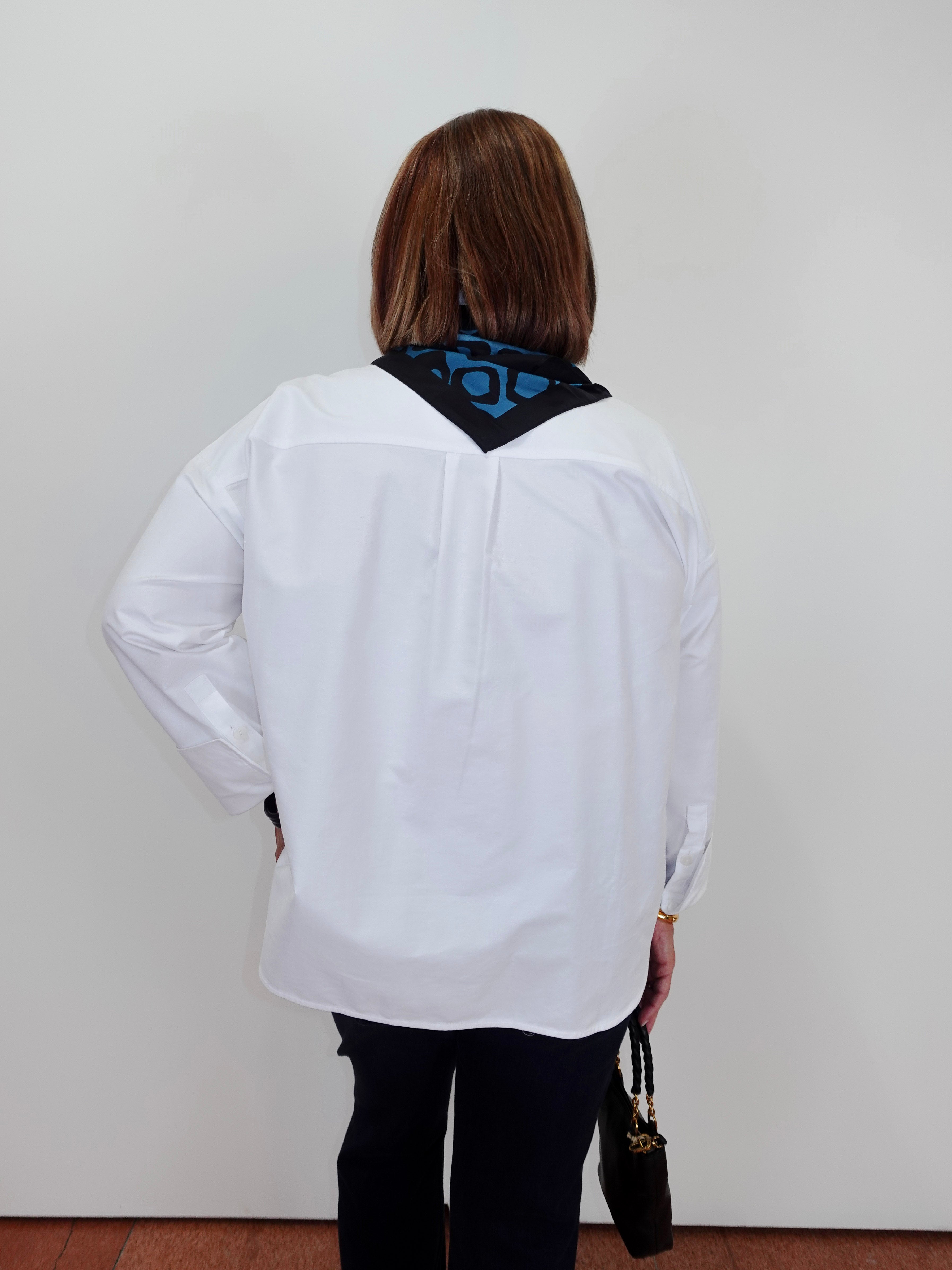 A woman viewed from behind wearing a white long-sleeve button-up shirt with a black neck scarf featuring a blue geometric pattern, paired with black pants and holding a small black handbag.
