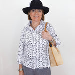 Woman wearing a black and white patterned shirt, gray pants, and a black hat, holding a beige handbag against a white background.