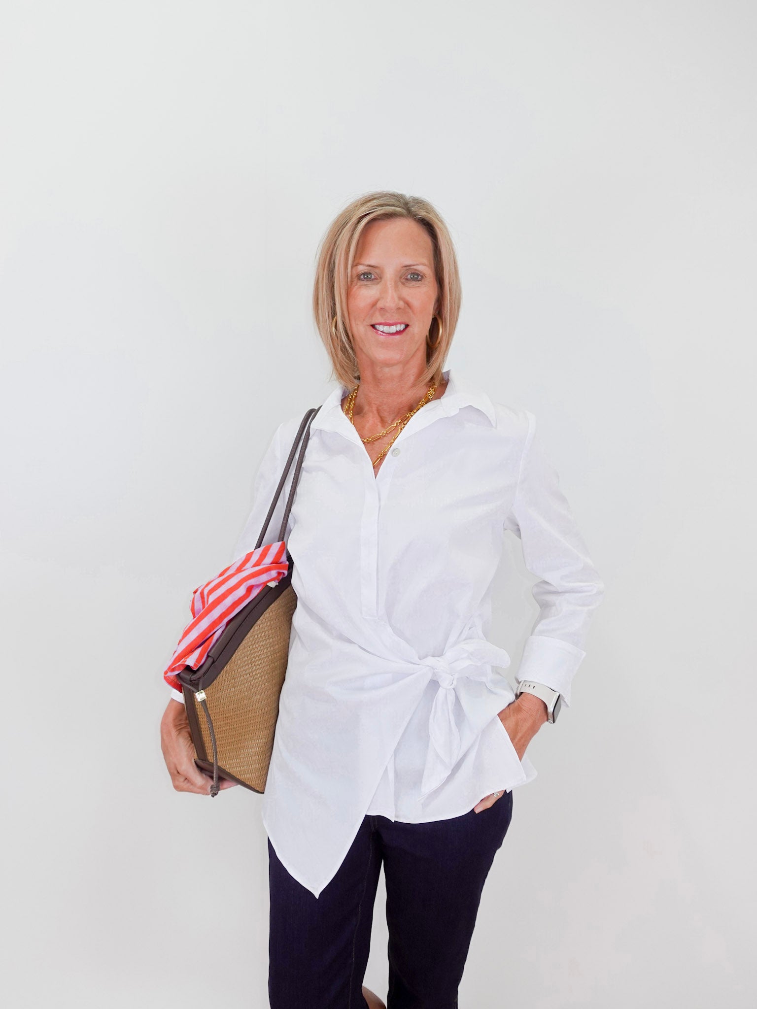 Woman wearing a white shirt and dark pants, holding a bag against a plain background