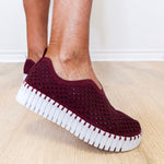 Maroon slip-on shoes with white sole worn by a person on a light wooden floor.
