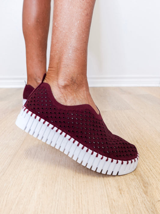 Maroon slip-on shoes with white sole worn by a person on a light wooden floor.