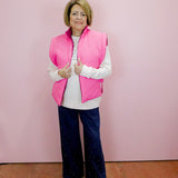 Woman wearing a pink vest over a white shirt with dark pants against a pink wall.