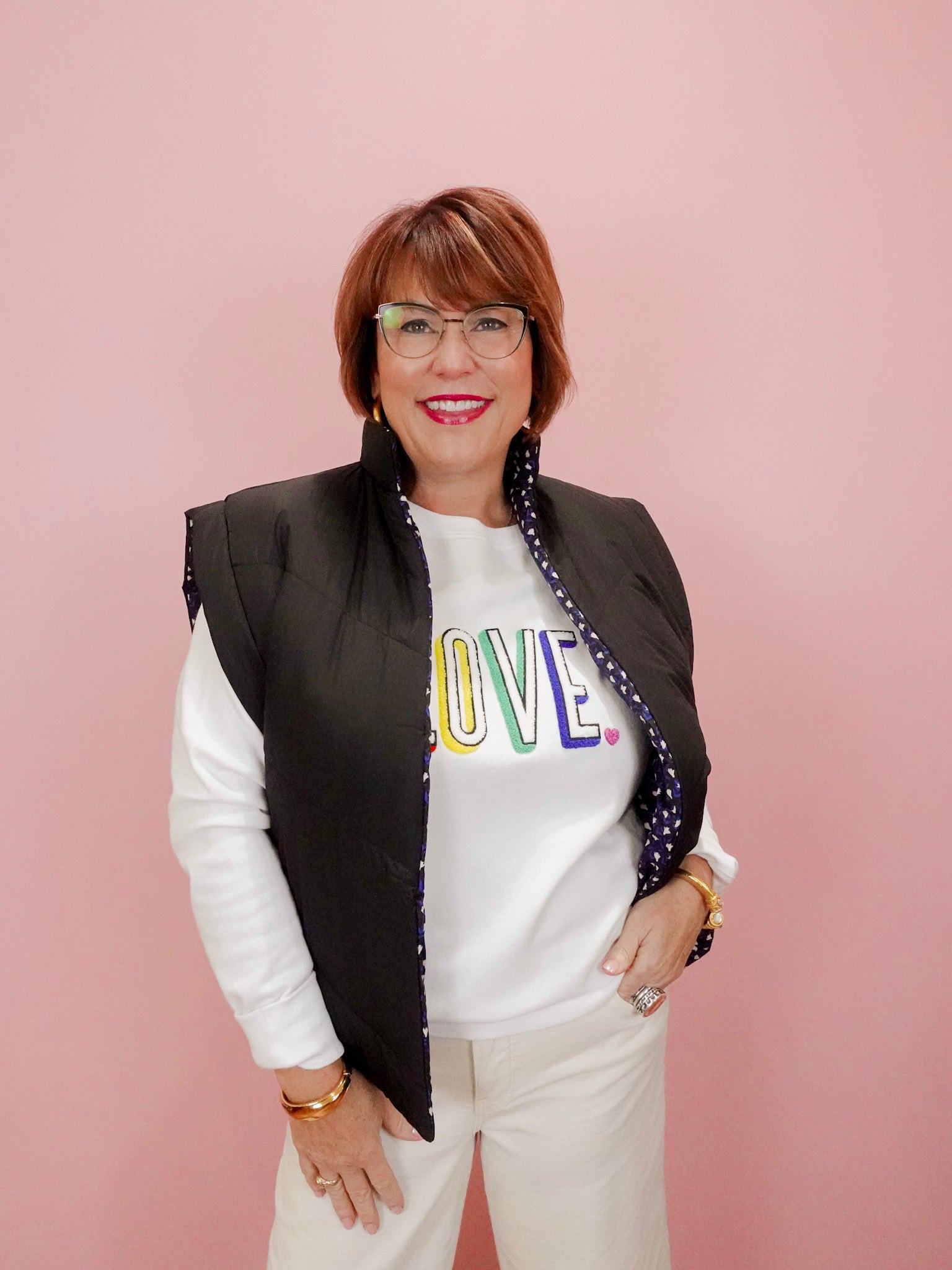 Woman wearing a white sweatshirt with colorful letters, black vest, and white pants against a pink background