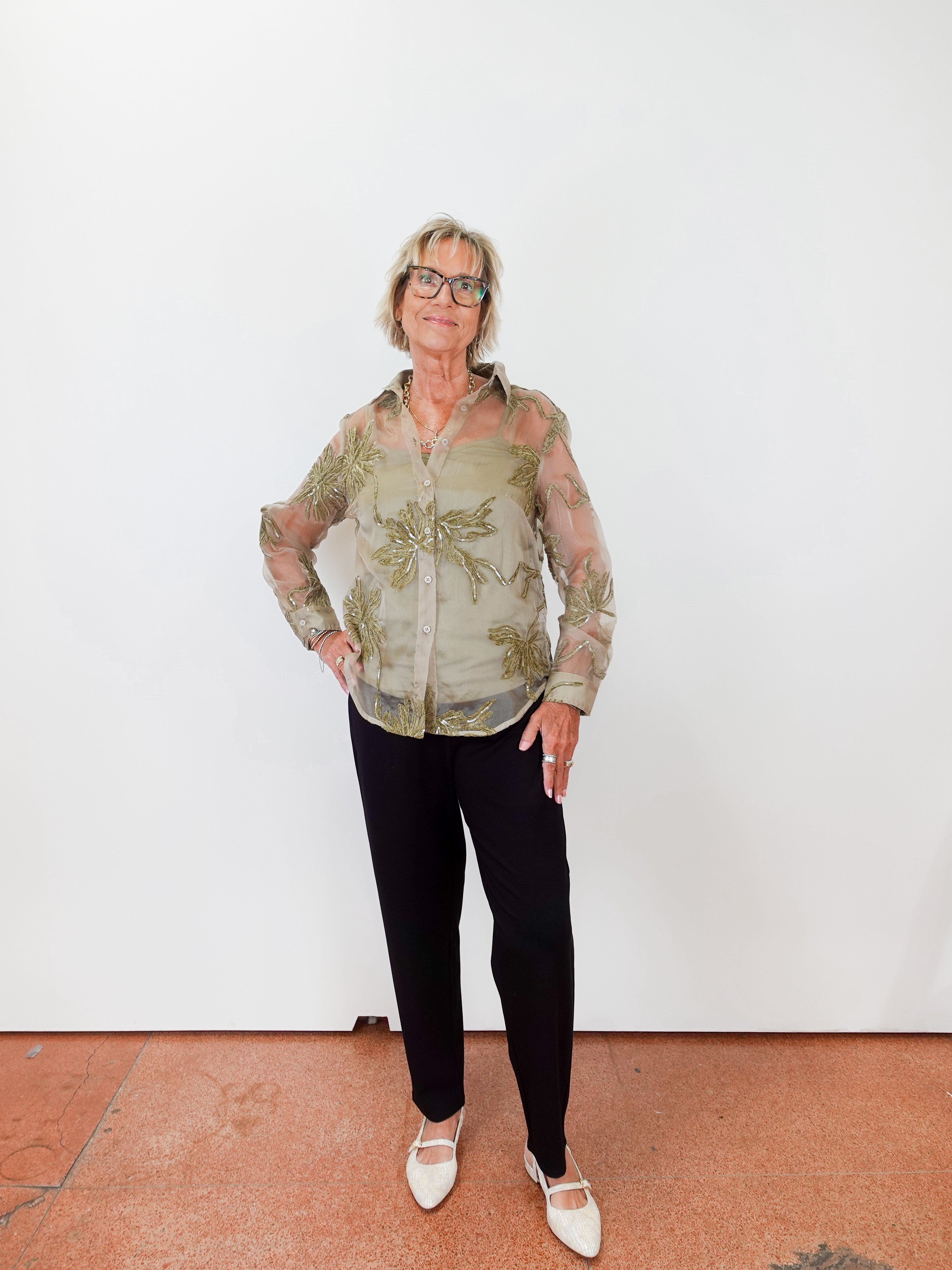 Person wearing a patterned shirt and black pants against a white wall.