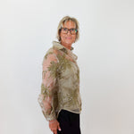 Woman wearing a sheer green floral blouse against a white wall.