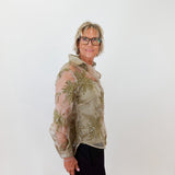 Woman wearing a sheer green floral blouse against a white wall.