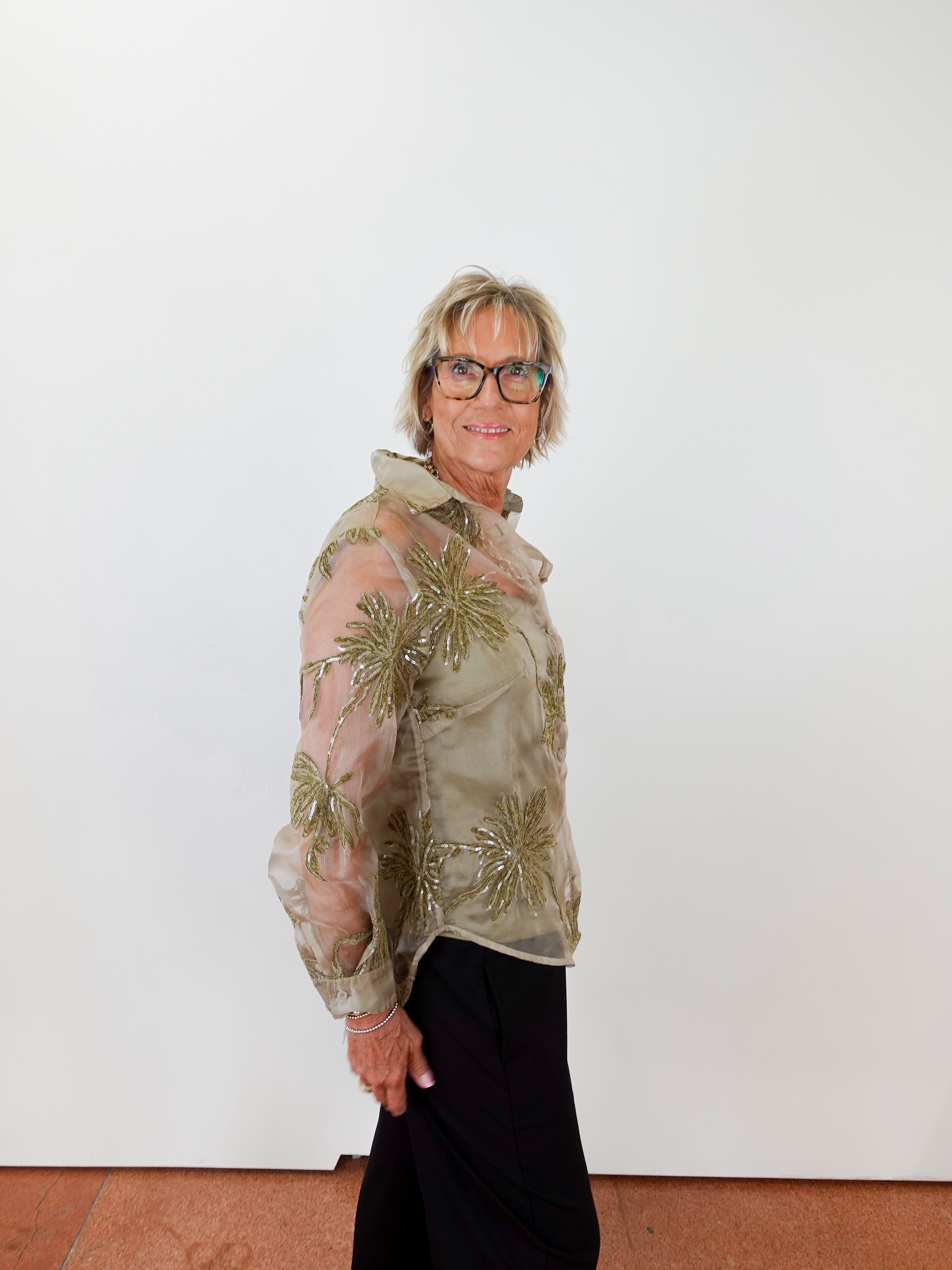 Woman wearing a sheer green floral blouse against a white wall.