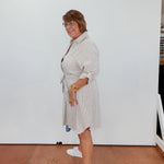 Woman wearing a light-colored dress with a belt, standing indoors against a white wall.