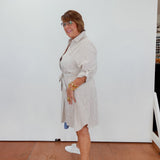 Woman wearing a light-colored dress with a belt, standing indoors against a white wall.