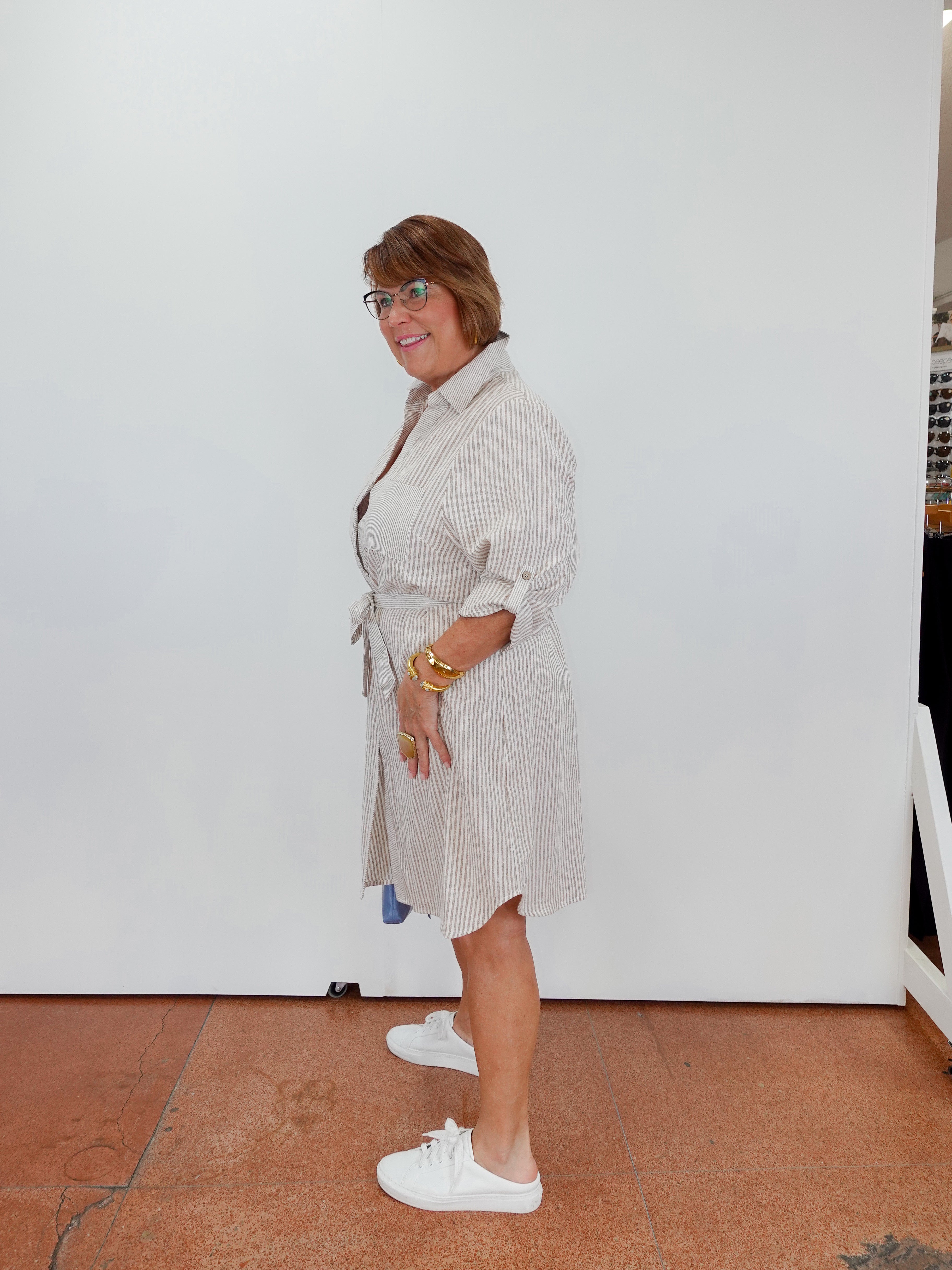 Woman wearing a light-colored dress with a belt, standing indoors against a white wall.