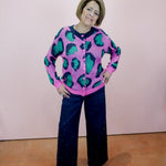 Person wearing a pink sweater with green leopard print and dark blue pants against a plain background