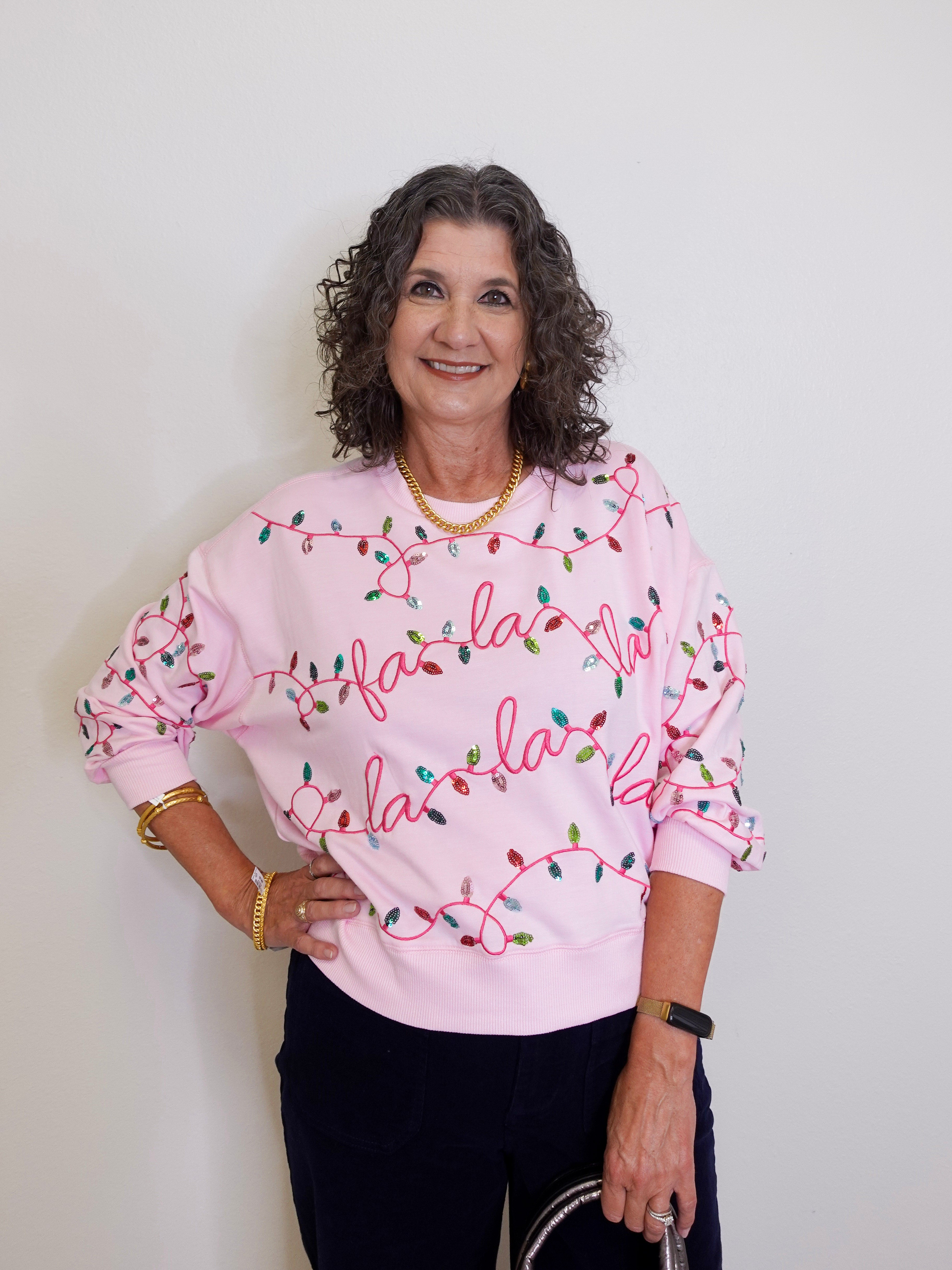Woman wearing a pink sweatshirt with 'fa la la' text and Christmas lights design against a plain background