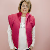 Woman wearing a pink vest over a white shirt with text, standing against a pink background