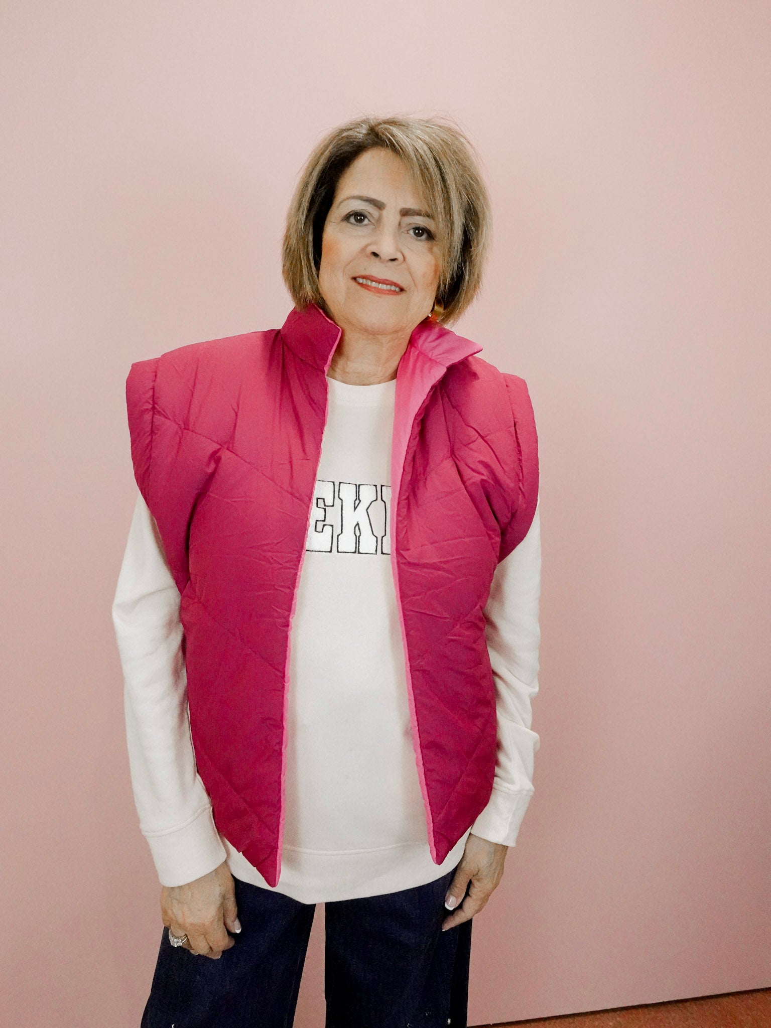 Woman wearing a pink vest over a white shirt with text, standing against a pink background