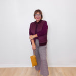 Woman wearing a purple jacket and plaid pants standing against a white wall.
