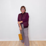 Woman wearing a purple jacket and plaid pants standing against a white wall.