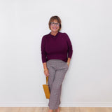 Woman wearing a purple sweater and patterned pants against a white wall.