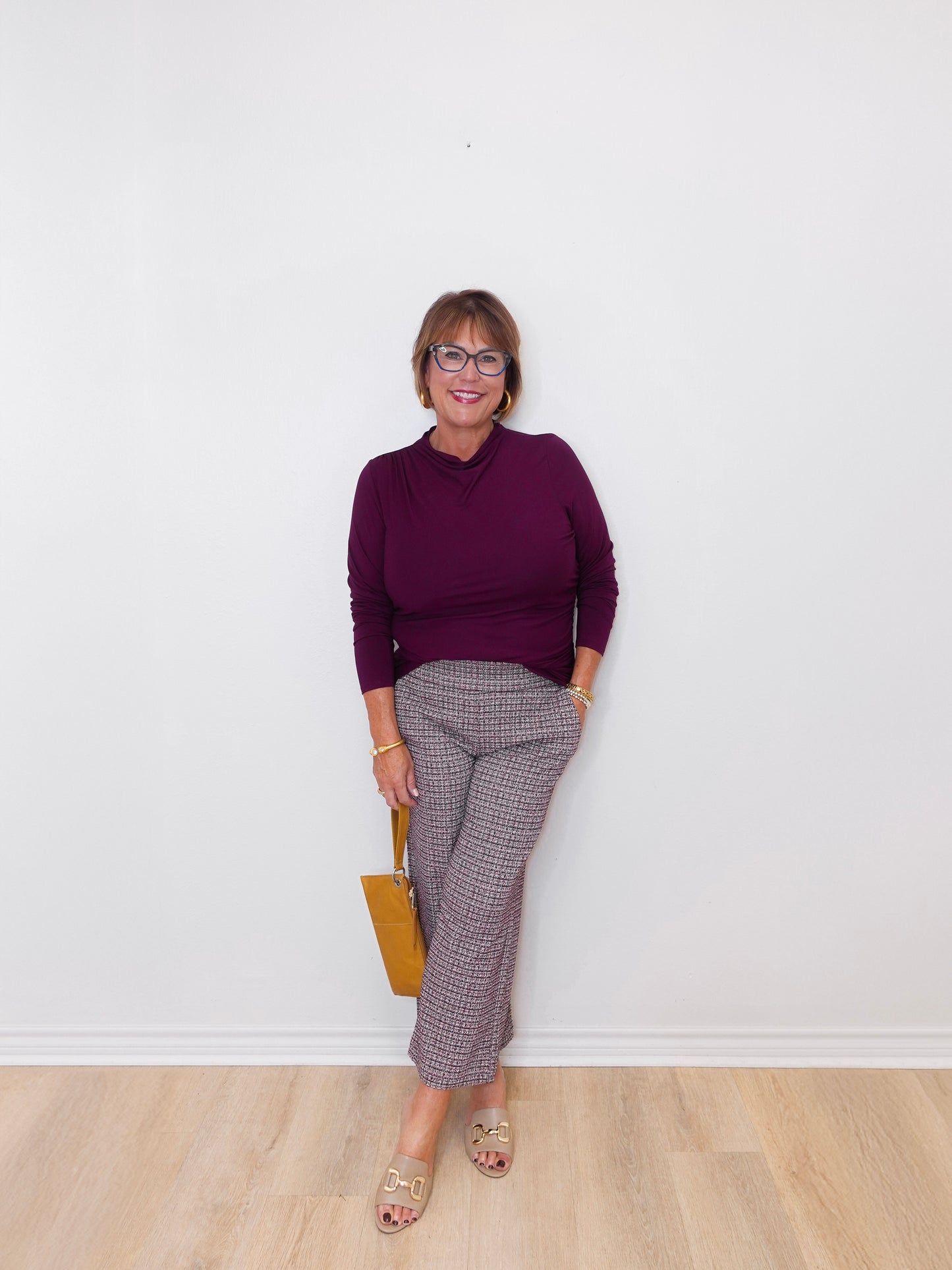 Woman wearing a purple sweater and patterned pants against a white wall.