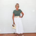 Woman wearing a green top and white skirt with a beige handbag against a white wall.
