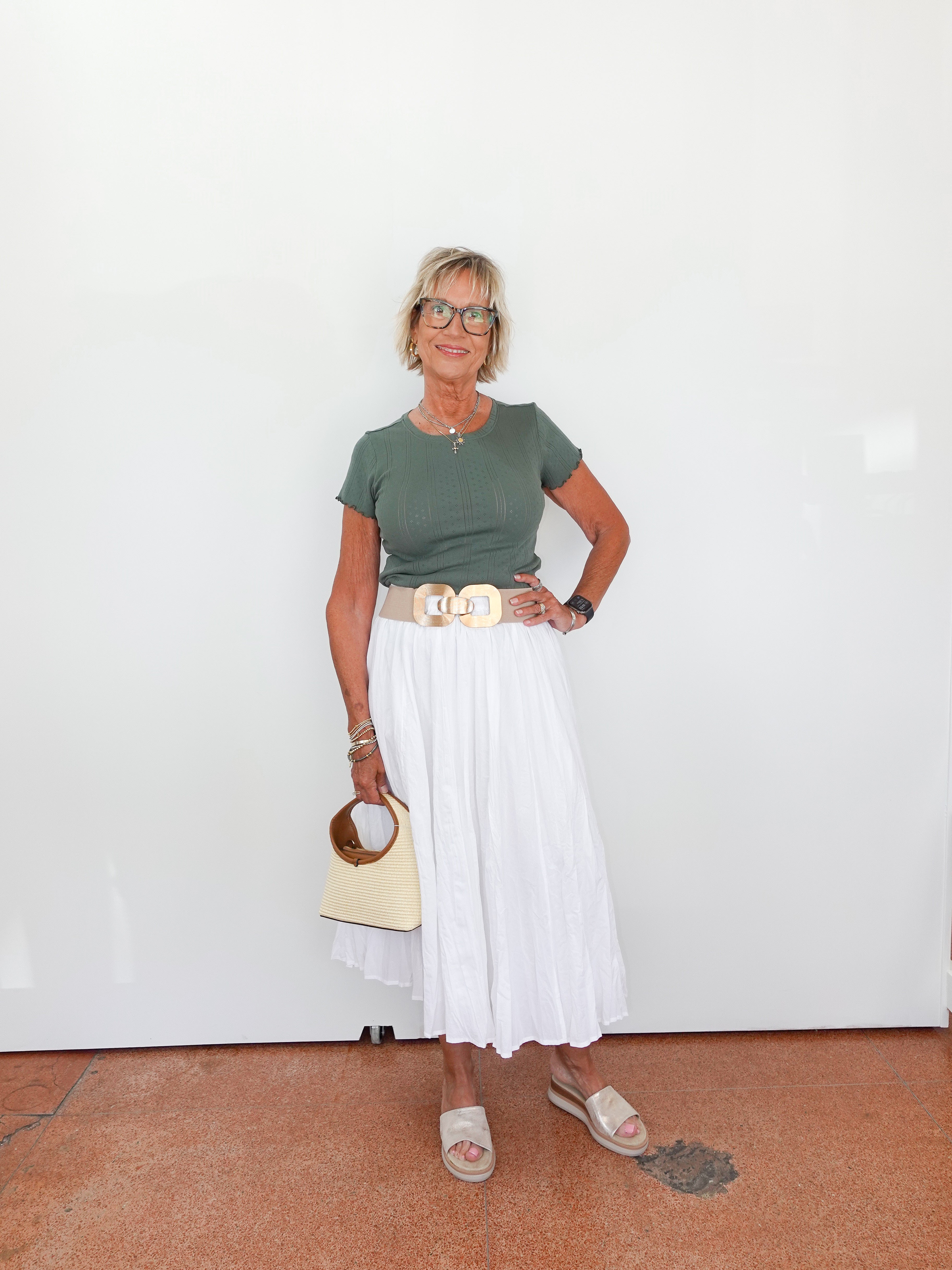 Woman wearing a green top and white skirt with a beige handbag against a white wall.