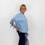 Woman wearing a blue polka dot top and dark pants against a white wall.