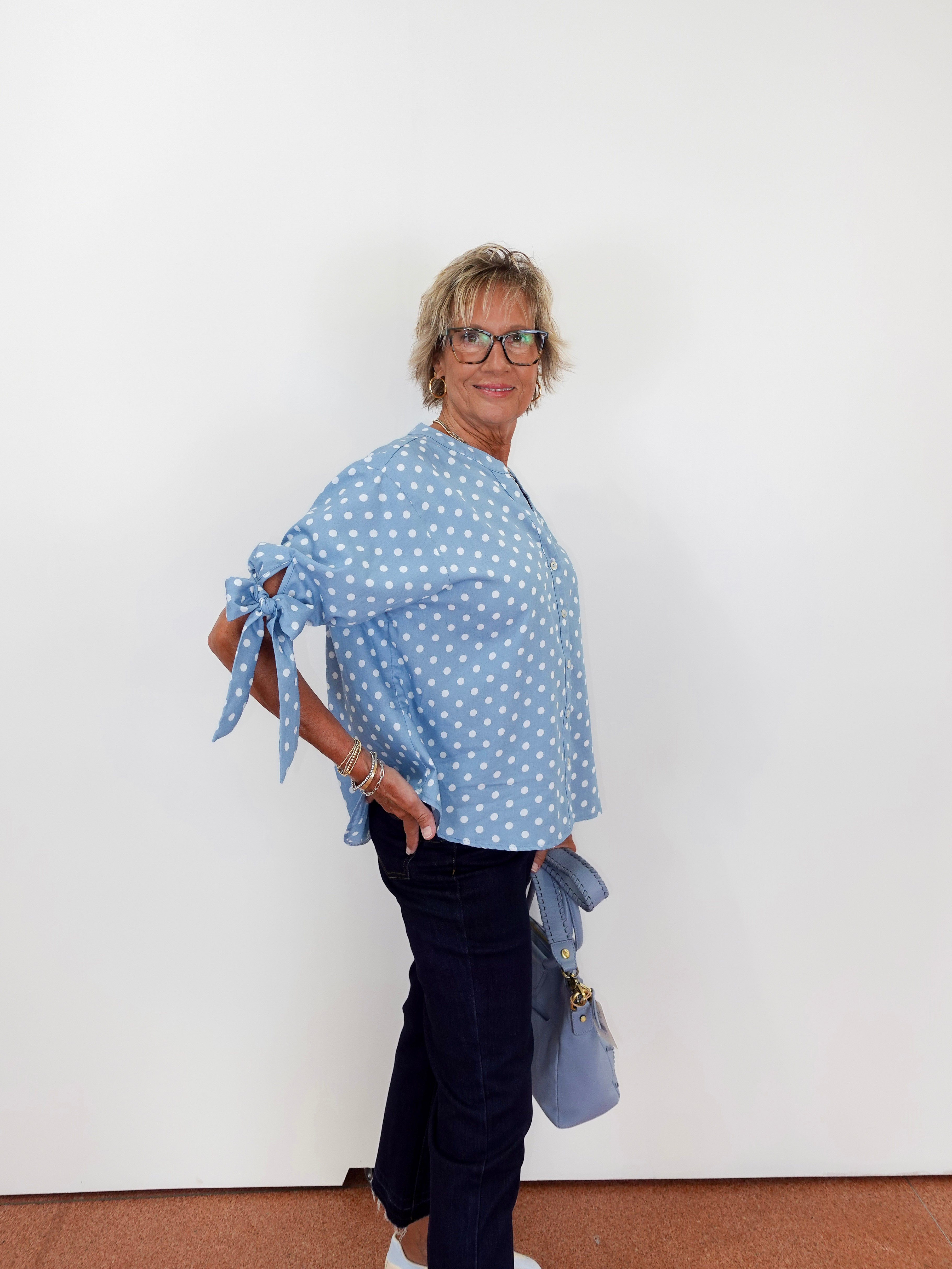 Woman wearing a blue polka dot top and dark pants against a white wall.
