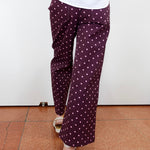 A person wearing dark purple wide-leg pants with a white polka dot pattern, paired with a white top and white heeled sandals, standing on a terracotta tiled floor against a white wall.