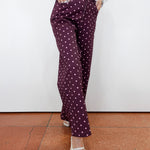 A person wearing dark purple wide-leg trousers with a white polka-dot pattern, styled with a white long-sleeve top and white pointed-toe flats, standing against a white wall and terracotta tiled floor.