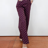 A person wearing dark purple wide-leg trousers with a white polka-dot pattern, styled with a white long-sleeve top and white pointed-toe flats, standing against a white wall and terracotta tiled floor.