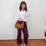 A woman stands against a white wall wearing a white button-up shirt, dark purple wide-leg pants with white polka dots, and white pointed flats. She is carrying a brown woven shoulder bag.