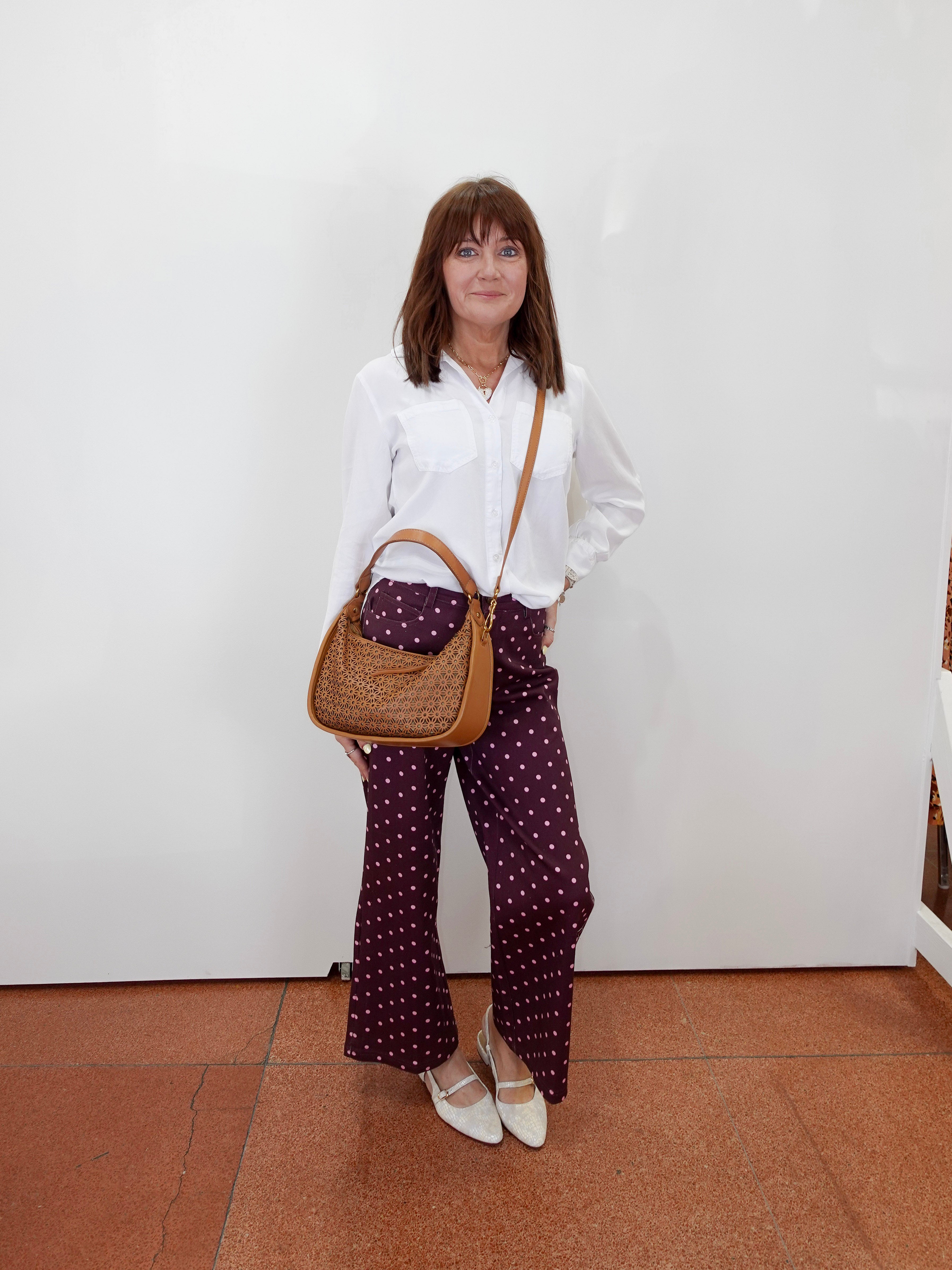 A woman stands against a white wall wearing a white button-up shirt, dark purple wide-leg pants with white polka dots, and white pointed flats. She is carrying a brown woven shoulder bag.