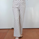 A person wearing white pants with a black polka dot pattern, a blue denim jacket, and tan sandals, standing against a white wall and terracotta tiled floor.