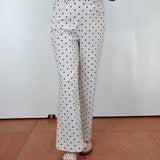 A person wearing white pants with a black polka dot pattern, a blue denim jacket, and tan sandals, standing against a white wall and terracotta tiled floor.