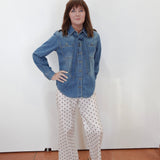 A woman stands against a plain white wall wearing a blue denim button-up shirt and white pants with black polka dots, paired with tan sandals.