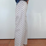 A person shown from the waist down wearing white wide-leg pants with a black polka dot pattern, styled with a blue denim jacket and brown sandals, standing against a white wall.