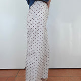 A person shown from the waist down wearing white wide-leg pants with a black polka dot pattern, styled with a blue denim jacket and brown sandals, standing against a white wall.