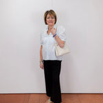 Woman wearing a white blouse and black pants holding a beige handbag against a white wall.