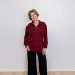 Woman wearing a red shirt and black pants standing against a white wall.