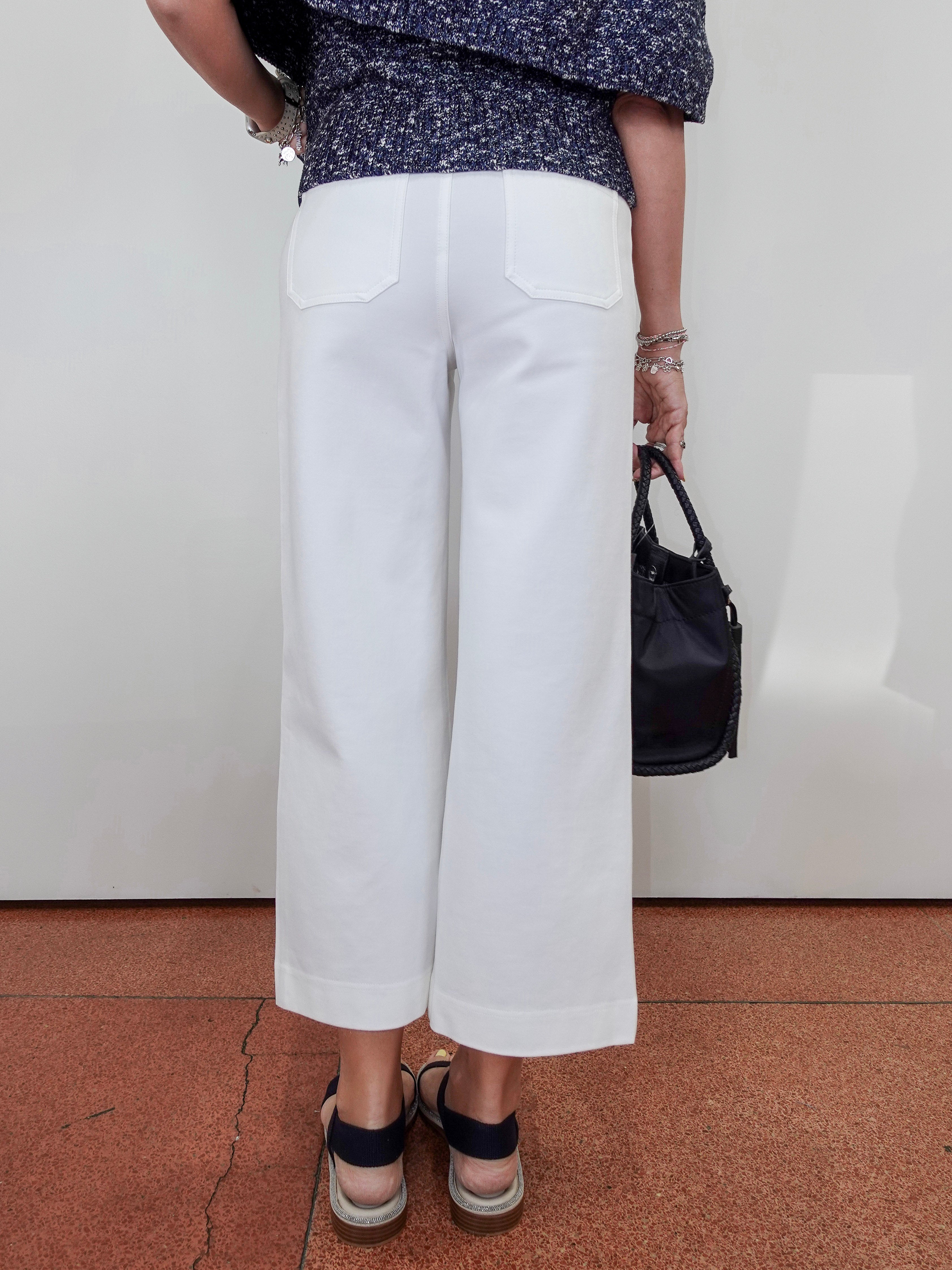 A person shown from the back wearing white wide-leg cropped pants, a navy patterned top, and black sandals, holding a black handbag against a plain light wall and terracotta floor.