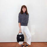 A woman stands against a white wall wearing a dark gray marled knit sweater, white wide-leg cropped pants, and black slide sandals, while holding a black handbag.