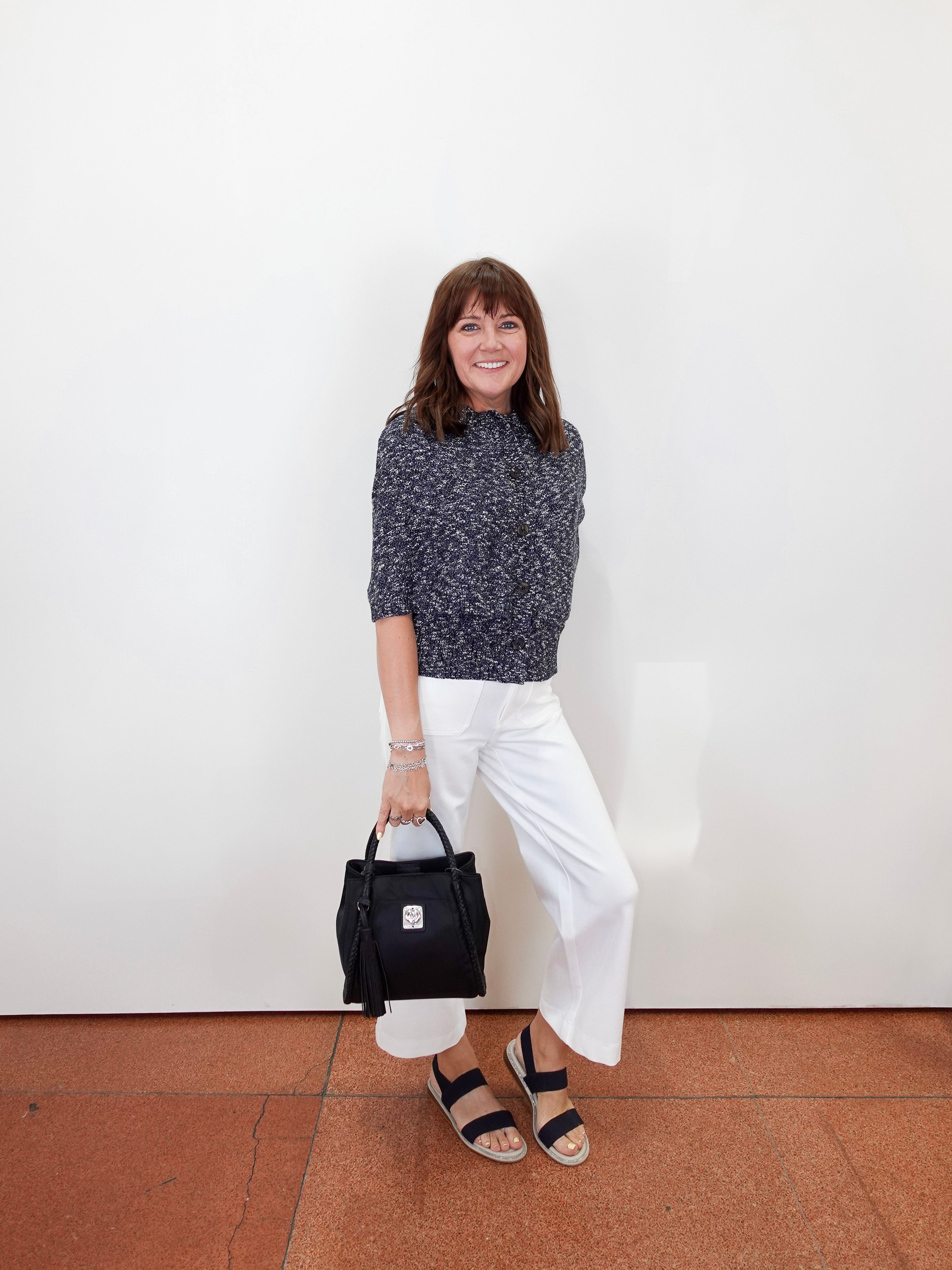 A woman stands against a white wall wearing a dark gray marled knit sweater, white wide-leg cropped pants, and black slide sandals, while holding a black handbag.
